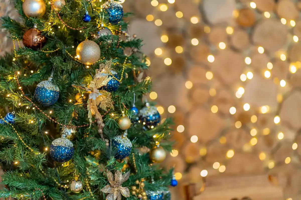 Árbol de Navidad decorado con esferas doradas y azules, luces y adornos dorados, con un fondo de luces desenfocadas.