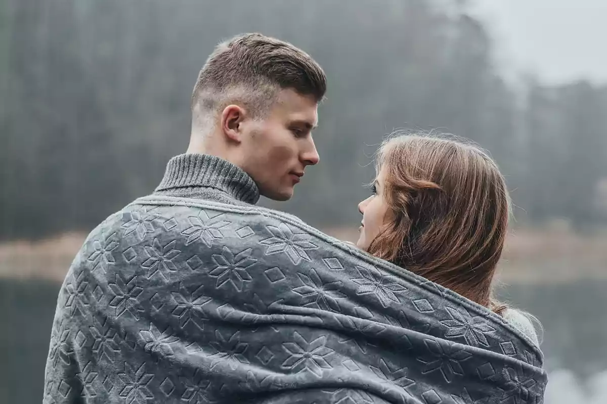 Una pareja envuelta en una manta gris con un diseño de copos de nieve, mirándose a los ojos en un entorno natural y nublado.