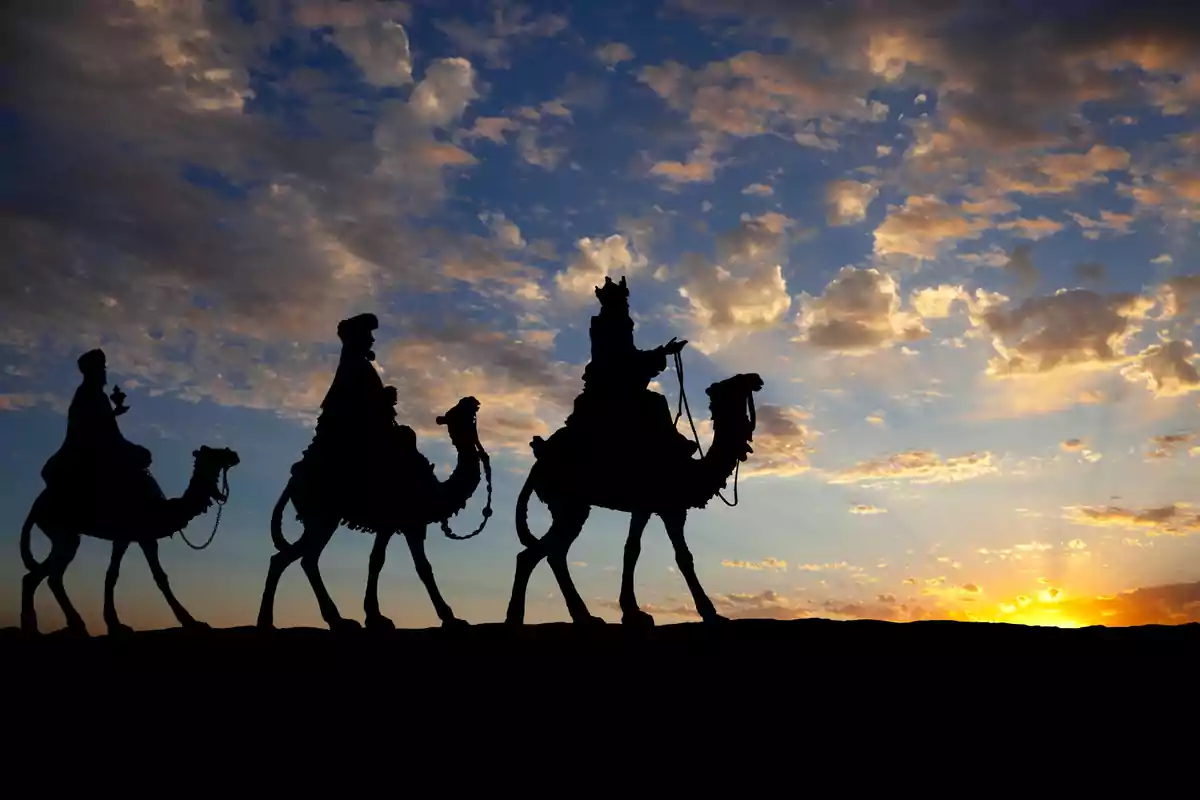 Tres siluetas de personas montando camellos al atardecer con un cielo parcialmente nublado.