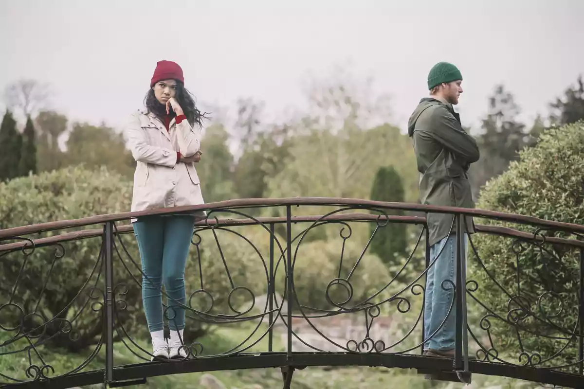 Una pareja de pie en un puente, ambos con los brazos cruzados y mirando en direcciones opuestas, con un fondo de vegetación y árboles.